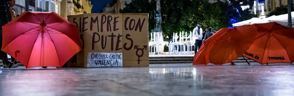 dos paraguas rojos rodeando una pancarta que lee "siempre con les putes. Orgull Crític València".