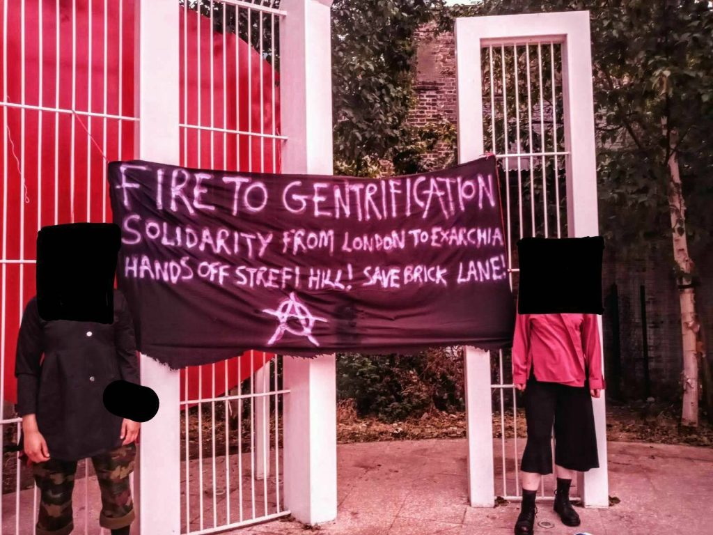 Photo of two people standing by the sides of a banner that is hanging in a park. The banner says "Fire to gentrification / Solidarity from London to Exarchia / Hands off strefi hill! Save Brick Lane!"