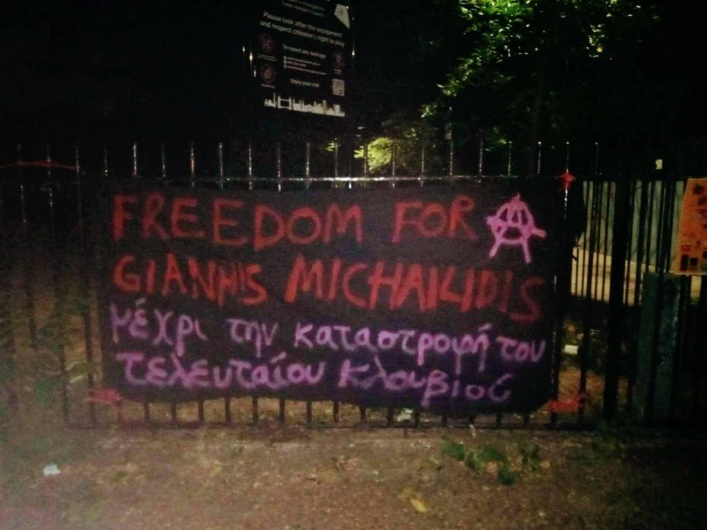 Photo of a banner hanging out in the fences of a park. The banner says "Freedom for Giannis Michailidis" in English and then again in Greek