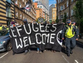 A large black banner reading "Refugees Welcome" with a half folded set of black and purple antifascist flags on one side is held by blurred figures, either side police officers wearing dark fatigues and hi vis vests flank the demonstration.