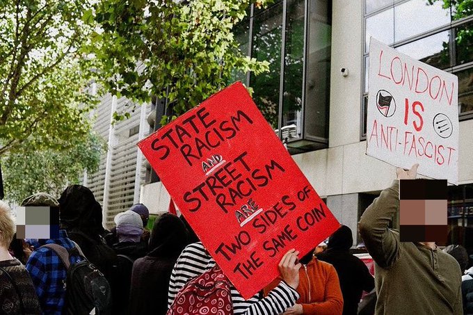 A person holds a red sign with black writing reading "State Racism and Street Racism are Two Sides of the Same Coin" with blurred figures at a demonstration around them.